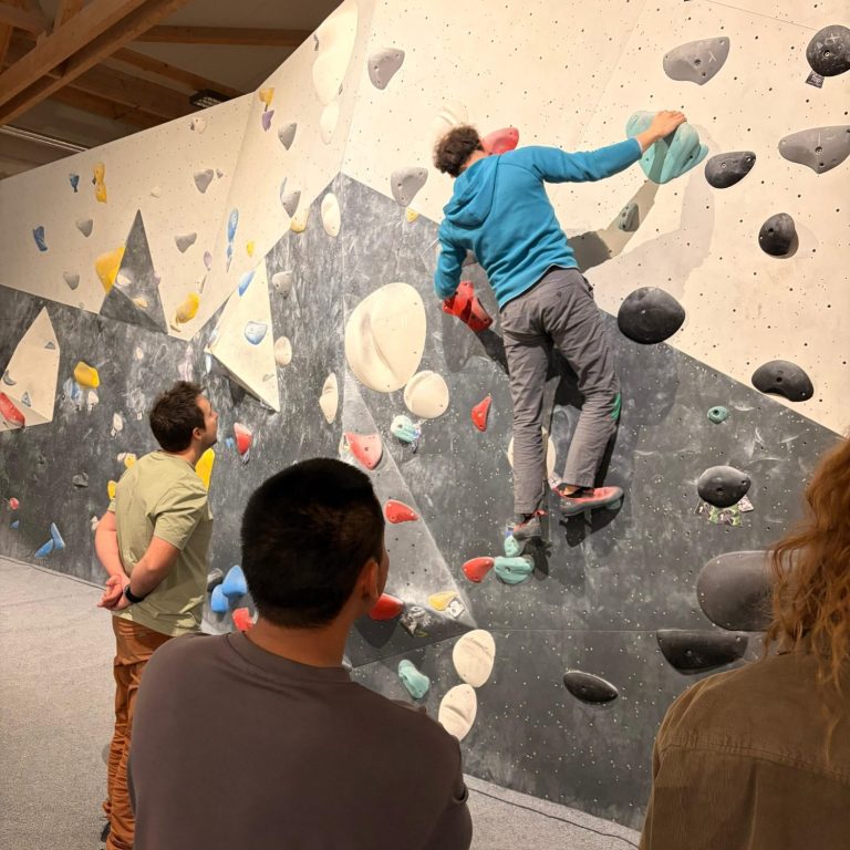 Gruppe beim Boulder-Teamevent in Düsseldorf: Eine Person klettert an der Boulderwand, während die anderen zuschauen und Tipps geben